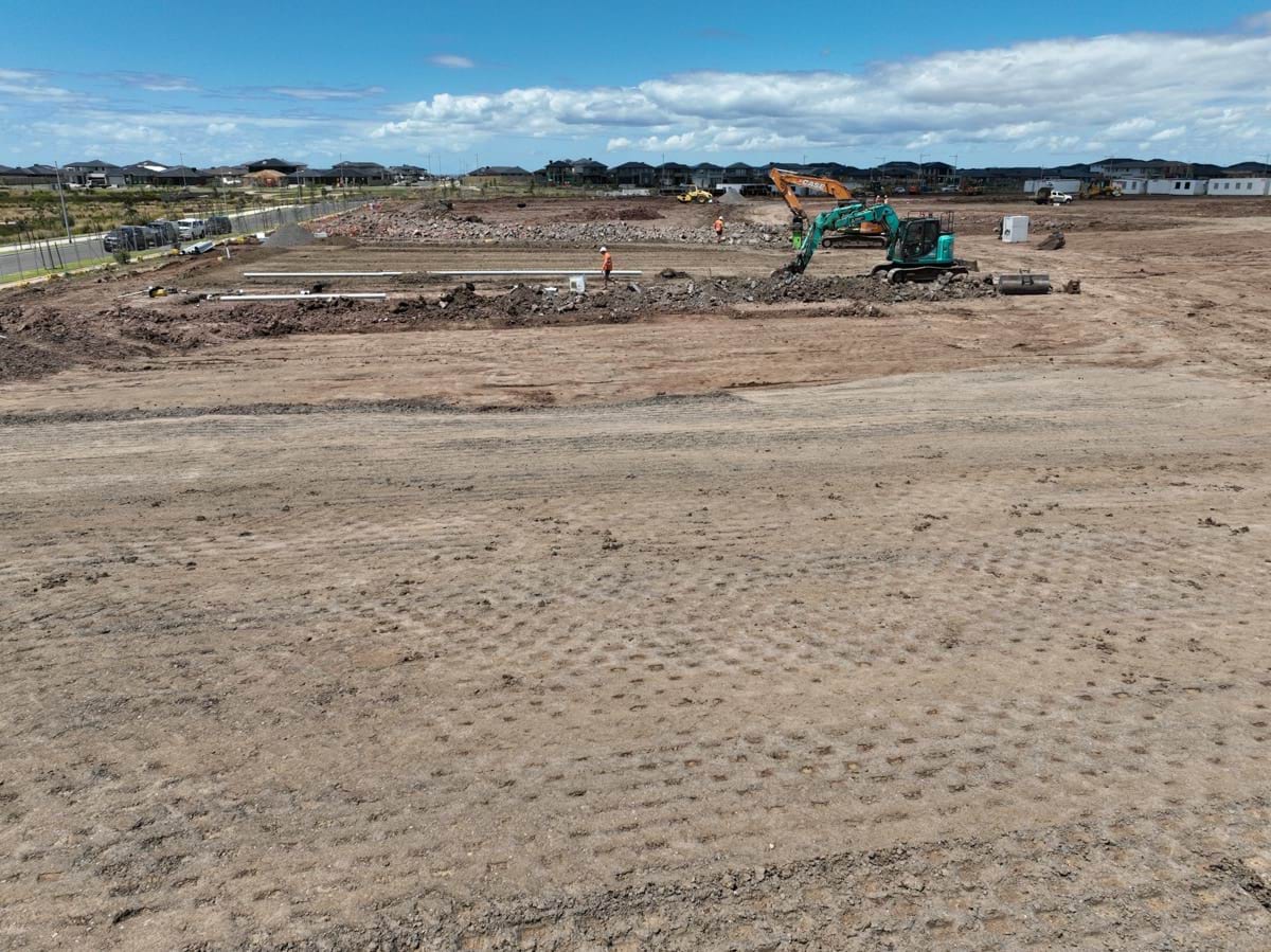 Black Forest East Primary School (interim name) - construction site progress in January 2023 - ground level shot showing flattened earth with a crane and a worker in the background.