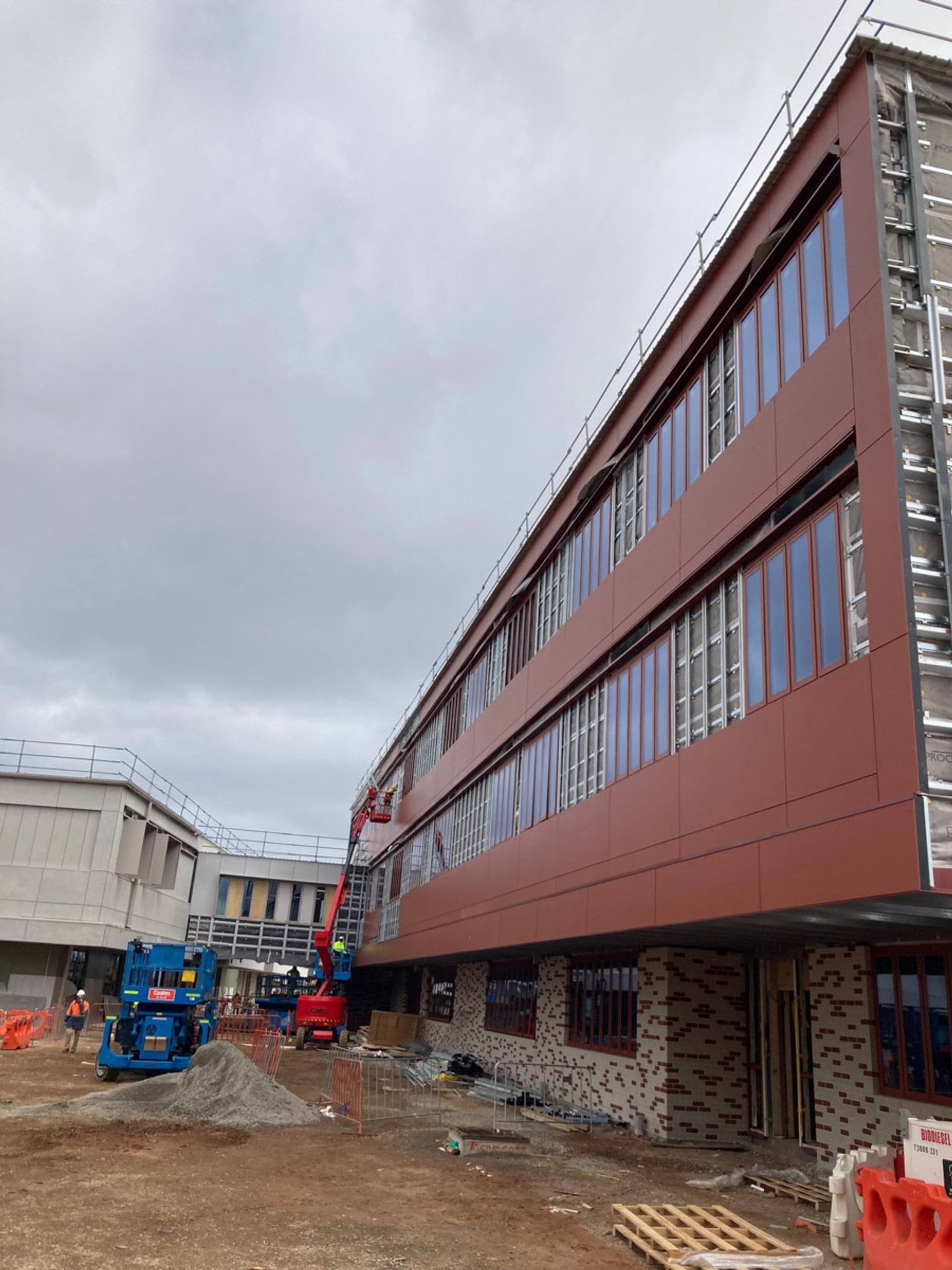 Exterior view of school building with red first and second levels and tiled ground level walls. Pallets, construction vehicles and unpaved earth surround this building