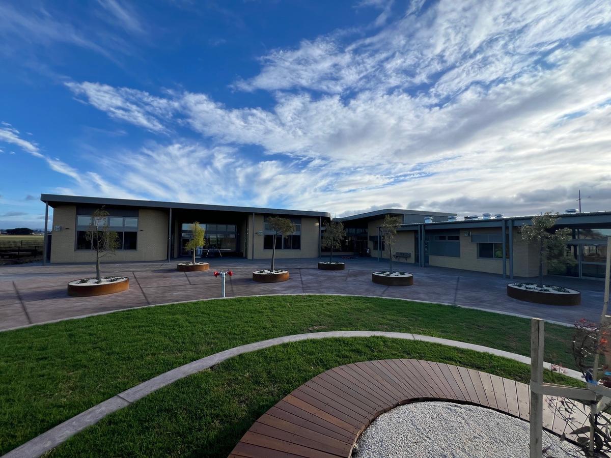 Raised garden beds have been placed in a zig zag pattern in a concreted area of the playground. Rounded landscaping features with grass wrapping around timber path