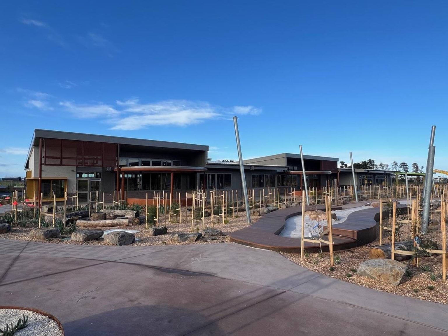 Playground landscaping features tanbark, sandpit surround by a timber path, and many short saplings and seedlings supported by timber stakes. The buildings in the background overlook playground.