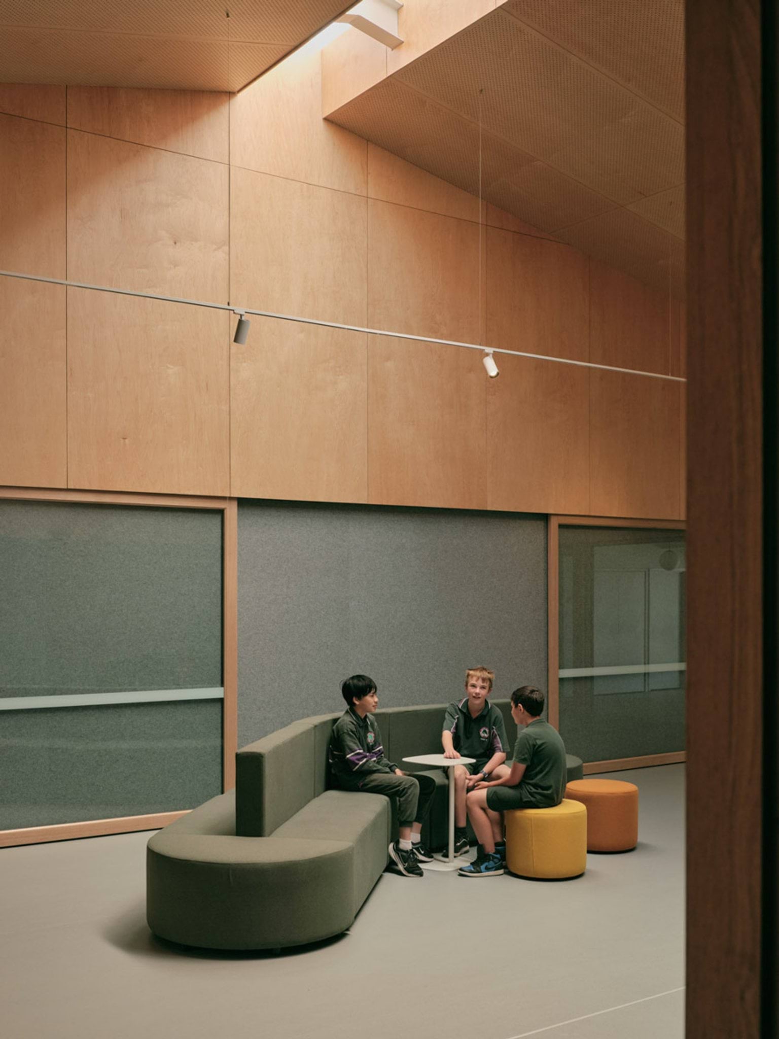 Students are sitting on a green couch and a yellow footstool around a small white table. The room they are in has a very high ceiling with a skylight. Behind them are sliding glass doors.