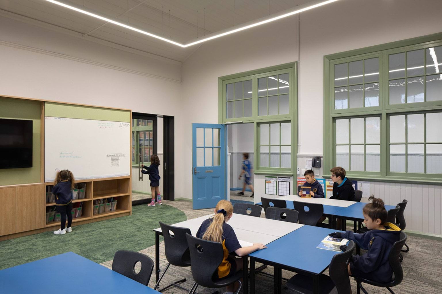 Inside a classroom at Moonee Ponds West Primary School. Kids are sitting at desks or standing near a whiteboard. There are big windows looking out into a hallway. 