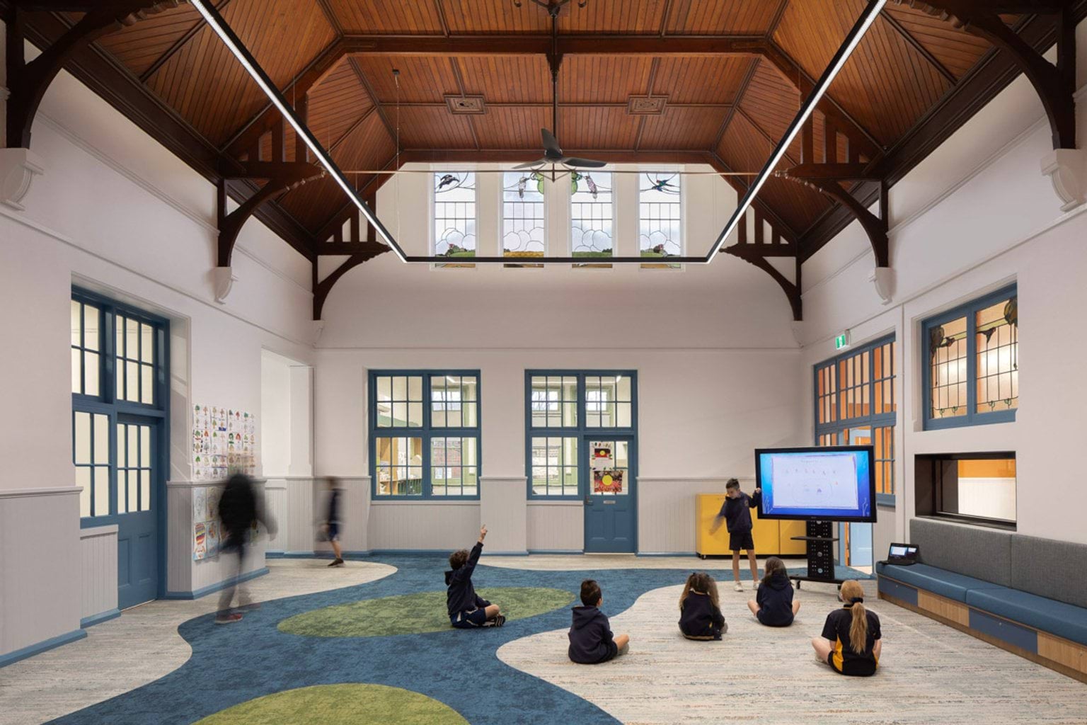 Inside a room at Moonee Ponds West Primary School. There is a very high wooden ceiling and white walls. There is a carpet with a colourful design. Kids are sitting on the carpet facing a big TV. There are lots of windows letting in light.