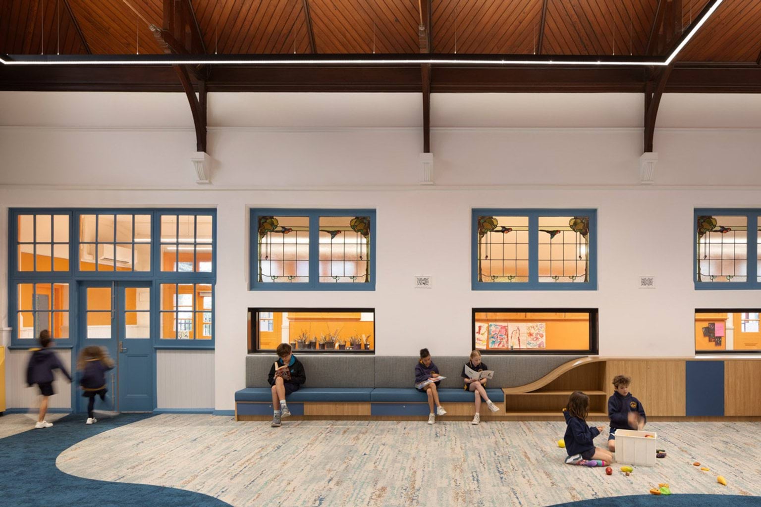 Inside a room at Moonee Ponds West Primary School. Children are moving around or sitting on a long bench seat reading, or sitting on the ground playing with a box of toys. The wall behind them has lots of windows facing out into a hallway. The ceiling is very high. 