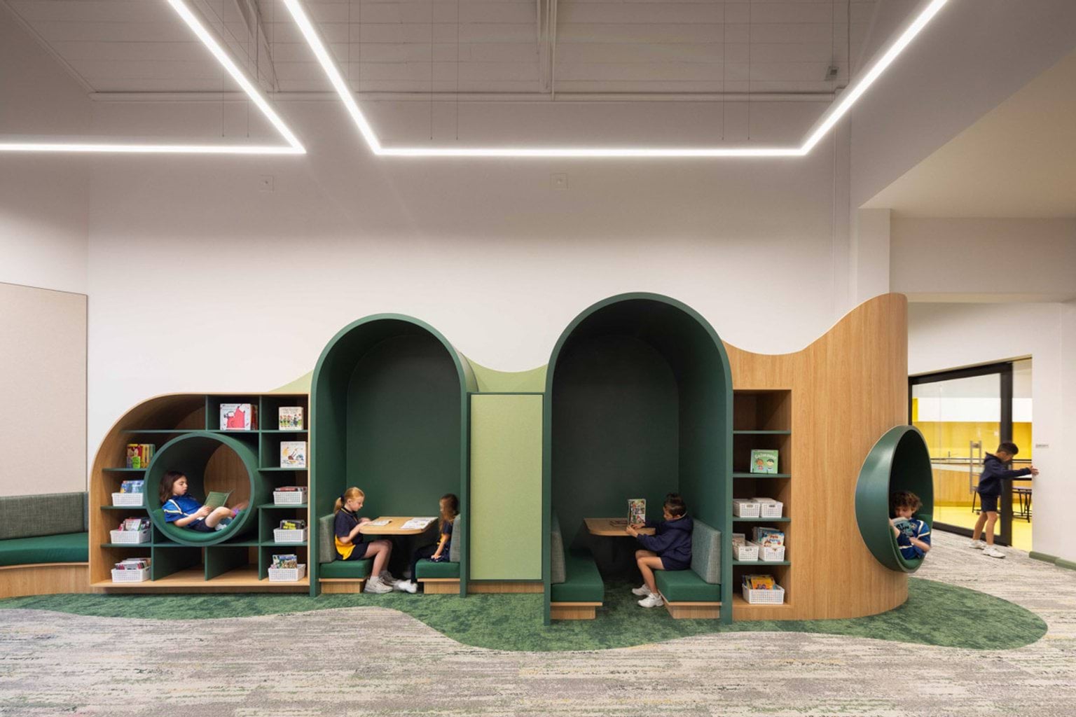 A wall at Moonee Ponds West Primary School that has recessed sections to sit in. 2 are circles where kids are sitting and reading, and 2 sections are arches with tables and bench seats. There are books and containers of equipment on shelving around these sections. 