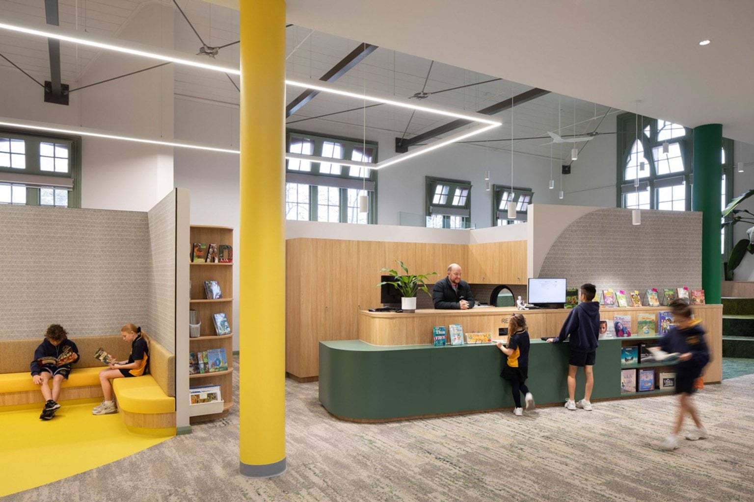 Inside what looks like the library at Moonee Ponds West Primary School. Kids are standing at a desk that an adult is behind, or sitting on bench seating reading. There are lots of books on shelves. A big yellow pole connects the ceiling to the ground in the middle of the photo. 