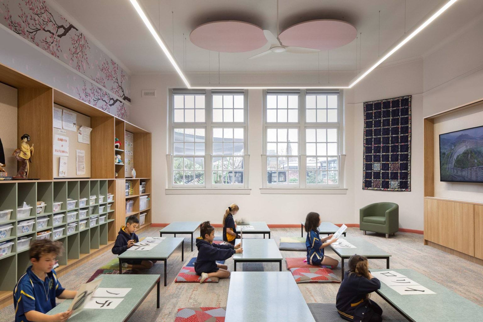Inside a Japanese language classroom at Moonee Ponds West Primary School. Kids are kneeling or sitting at low tables. They are looking at pages with Japanese language characters printed on them. There is shelving on the left and big windows at the back. There is wallpaper with Japanese cherry blossom trees. 
