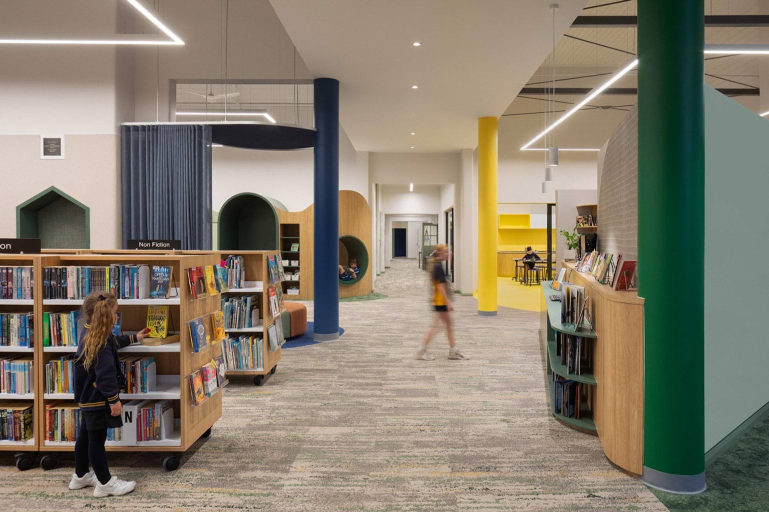 Inside a library at Moonee Ponds West Primary School. There are rows of low bookshelves filled with books. A student is standing at one of them. There are coloured poles connecting the ceiling and ground: blue, yellow and green. 
