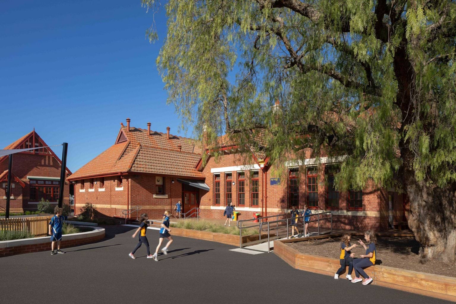 Outside Moonee Ponds West Primary School. Kids are running around or sitting together and playing. The school building  has a red brick exterior. There is a big tree with leaves in the right of the photo. 