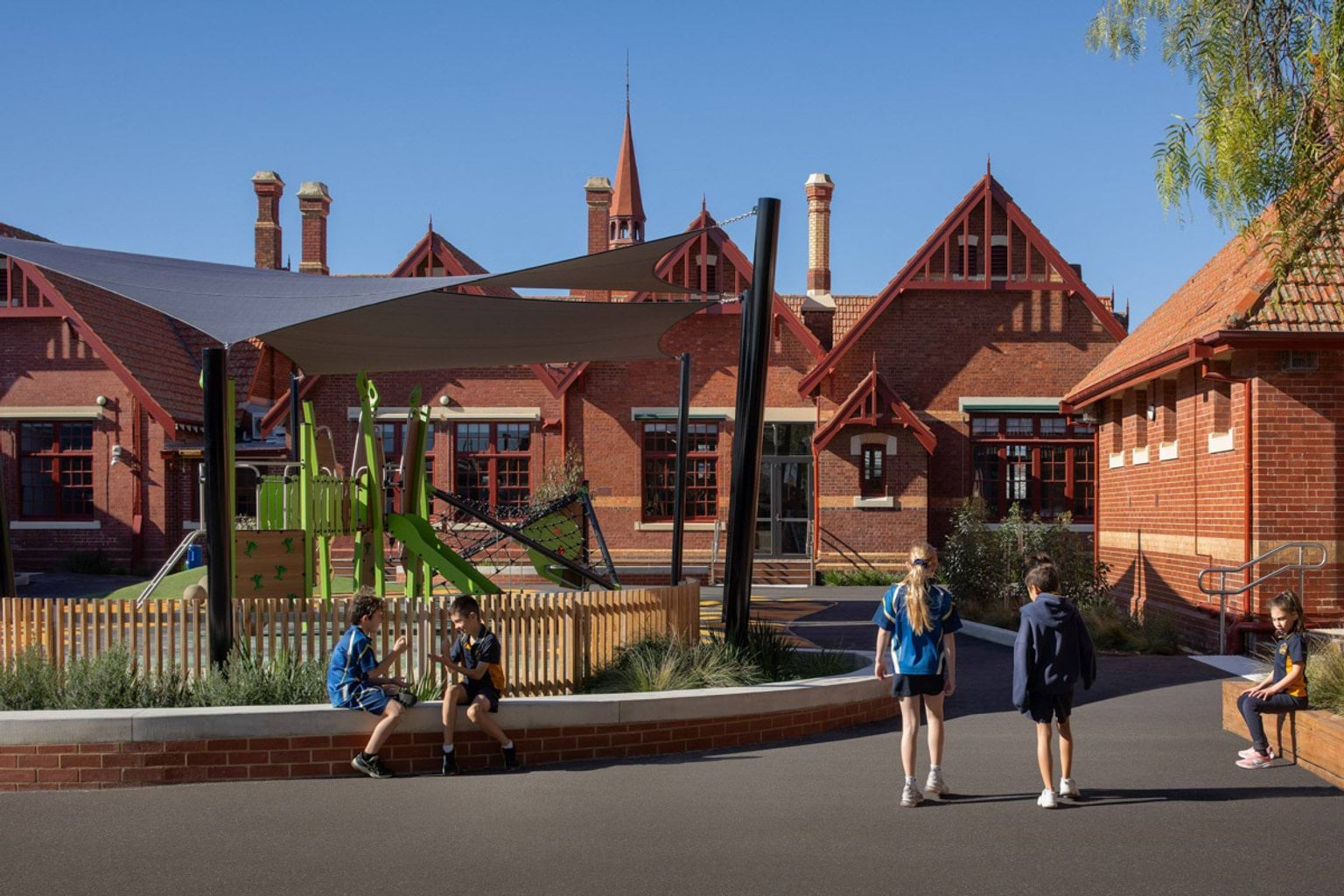 Outside Moonee Ponds West Primary School. The building has a red brick exterior. The playground is in front of it covered by shade sails. Kids are sitting down playing or walking past. 