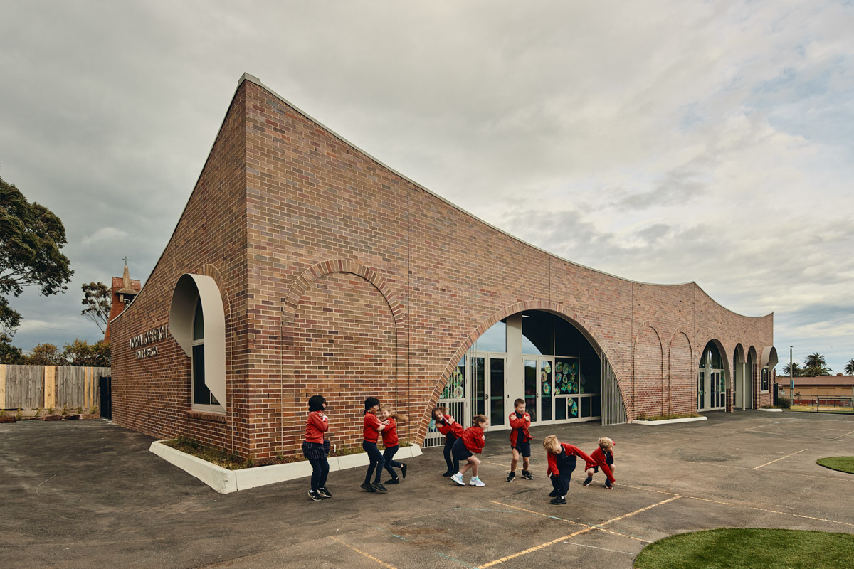 Outdoor photo of school administration building. It is a brown brick building with arches above its doors and windows. There is a group of children in school uniforms standing outside.