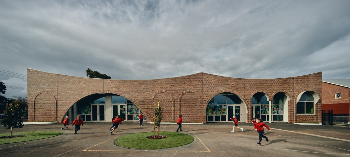 Outdoor photo of brick administration building. There are arches above the doors and windows, a concrete area outside, and saplings planted in grass circles. Children in school uniforms are walking in front of the building.