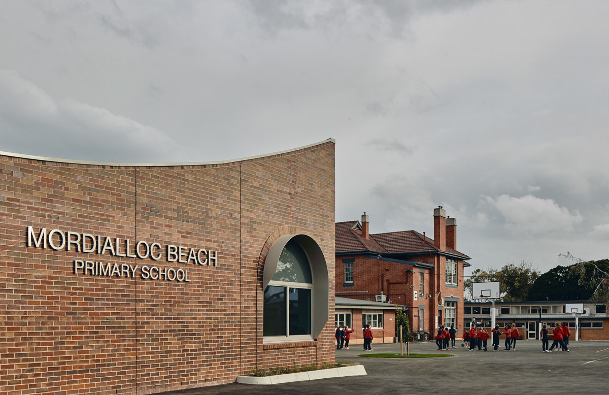 Outdoor photo of brick administration building. There are arches above the doors and windows, a concrete area outside, and saplings planted in grass circles. Children in school uniforms are walking in front of the building.
