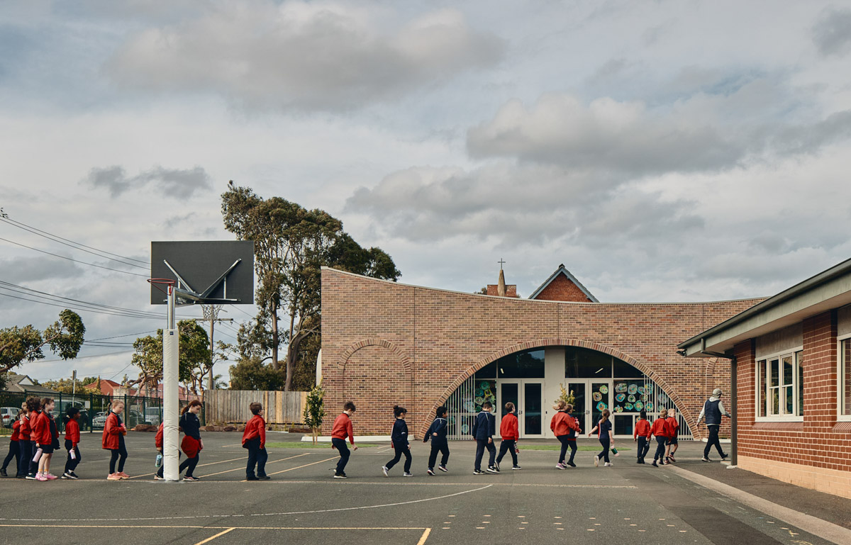 Outdoor photo of brick administration building. There are arches above the doors and windows, a concrete area outside, and saplings planted in grass circles. Children in school uniforms are walking in front of the building.
