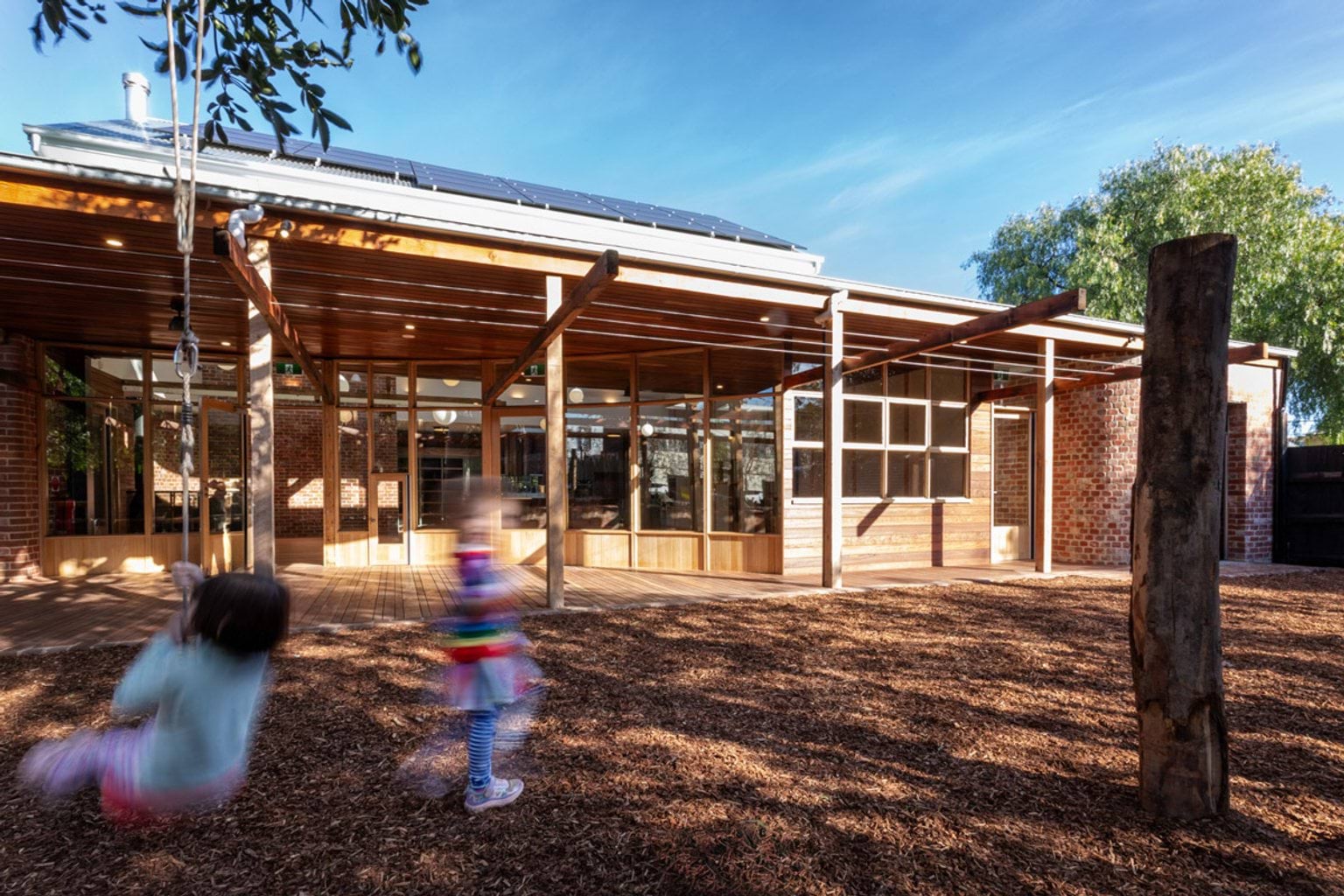 The outside of St Kilda & Balaclava Kindergarten, with 2 little kids blurred in the foreground. The building is made of wood with solar panels on the roof, and the ground in front is bark chips.