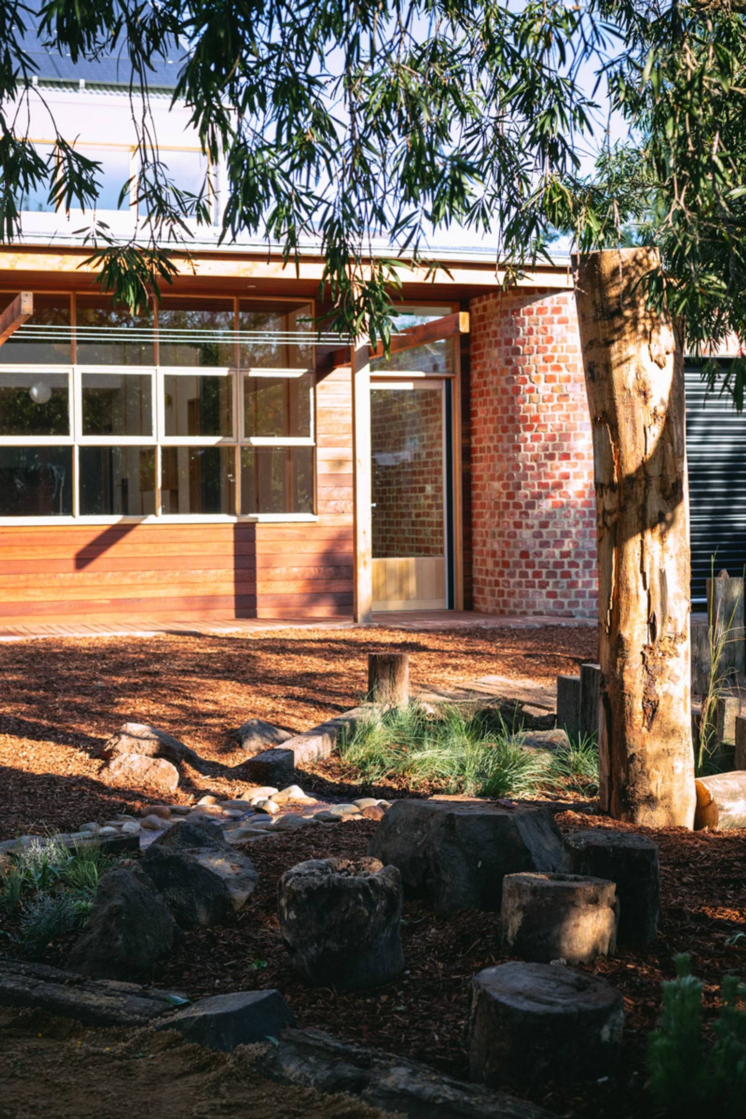Outside of St Kilda & Balaclava Kindergarten. The building is in the background, and there is a landscaped area in the foreground along with a tree stump and branches. 