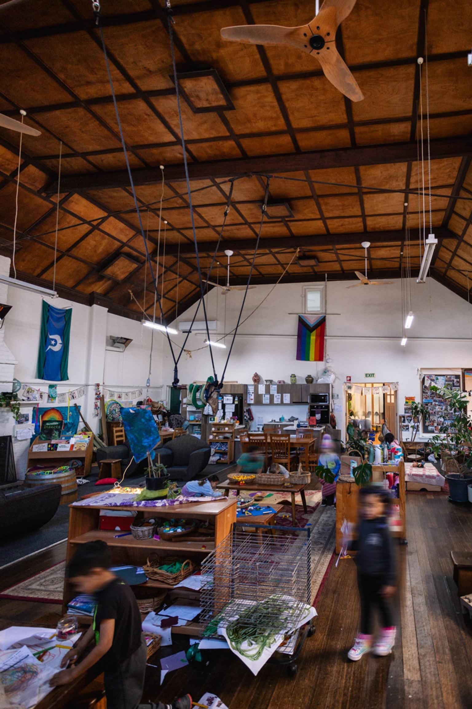 Inside St Kilda & Balaclava Kindergarten. It is a large room with high ceilings, white walls and a brown ceiling with lights hanging from it. The room is full of furniture and little kids moving around. A rainbow flag and Torres Strait Island flag hang from the walls.