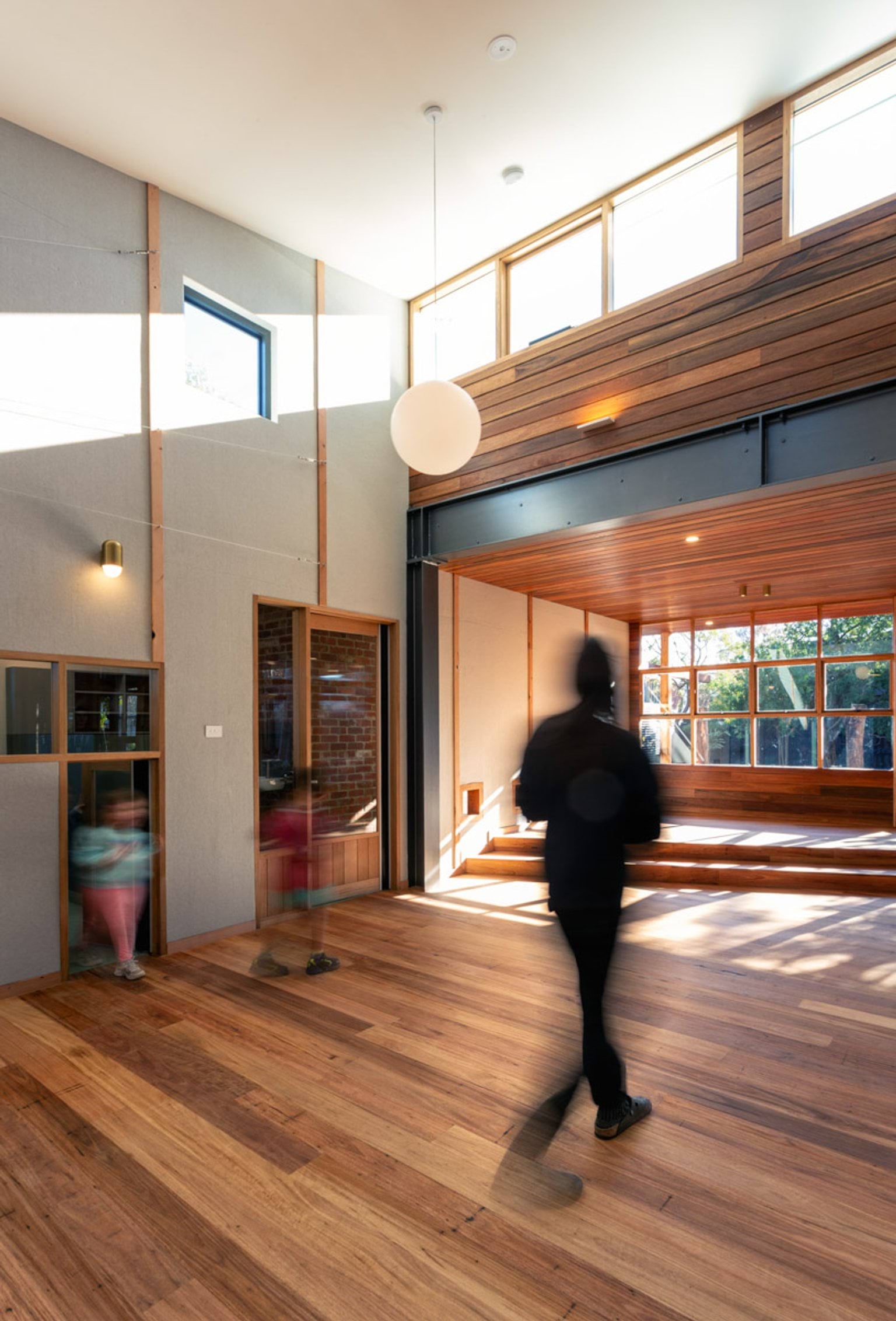 Inside St Kilda & Balaclava Kindergarten. There are 2 little kids and an adult walking. The floor is wooden and the ceilings are high. There are lots of windows letting in lots of sunlight. 