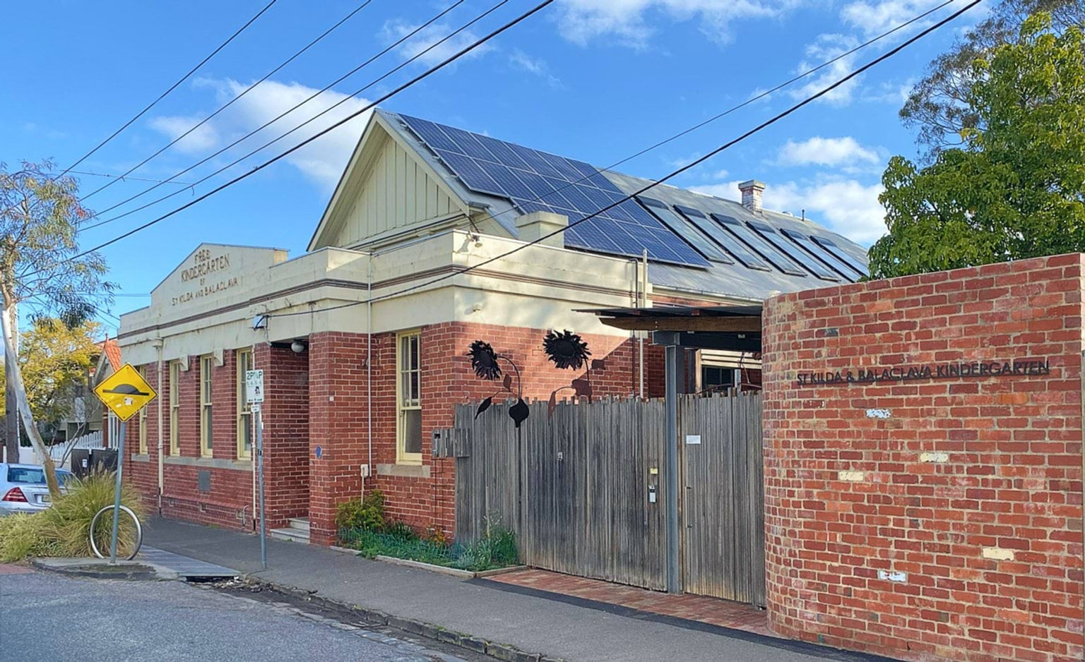 Outside St Kilda & Balaclava Kindergarten, looking at the building from a street. The outside has brick walls, with the kindergarten's name as a sign on a wall. There are plants hanging over a wooden fence. 