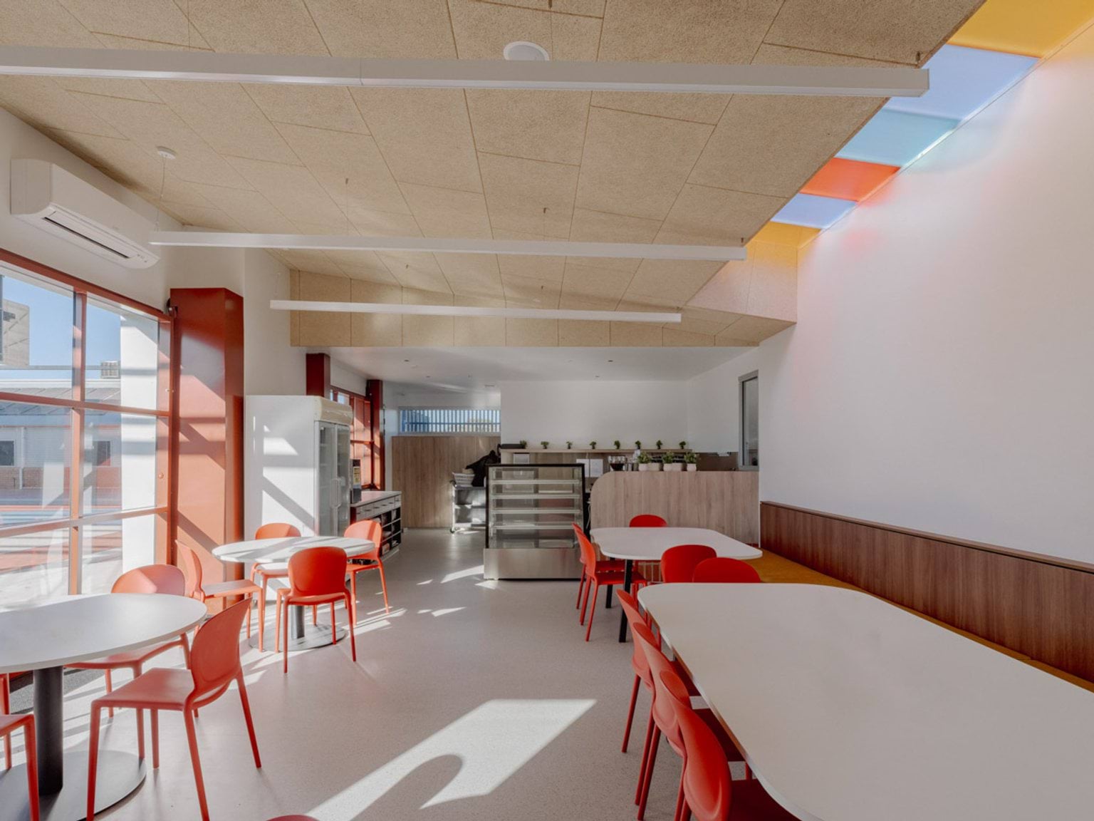 Inside the dining area of Swan Hill Specialist School. There are long tables with red chairs  and a small kitchen area with a fridge at the back of the room. There are pastel coloured glass skylights in a row in the ceiling.