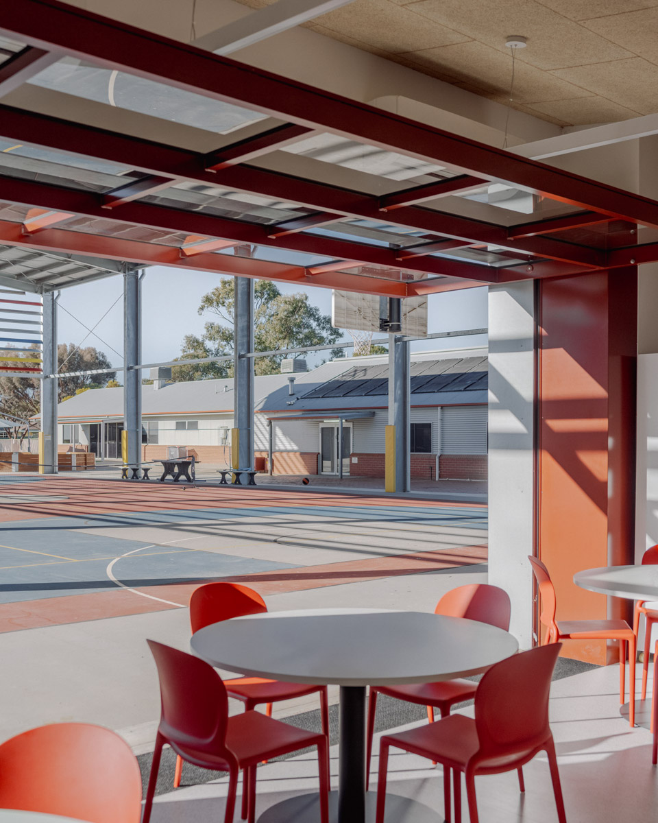 Standing inside the dining area at Swan Hill Specialist School and looking outside to the hardcourts. There is a table with red chairs in the foreground, and a glass panel of windows that has been swung open like a garage door to provide access between inside and outside. 