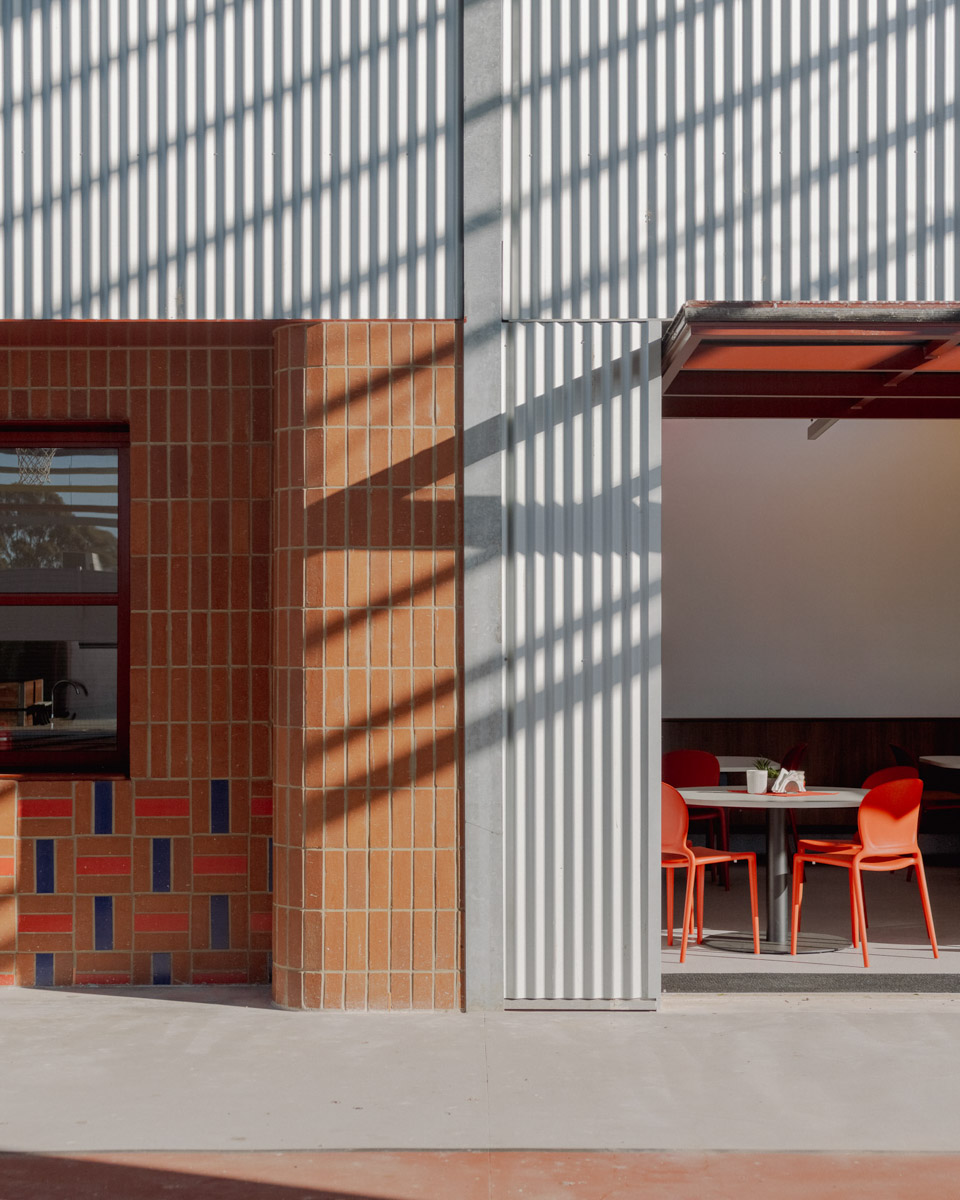 A close-up of the outside of a Swan Hill Specialist School building, showing a brick wall on the left next to a metal-clad wall on the right. The dining area with tables and red chairs is visible inside. 