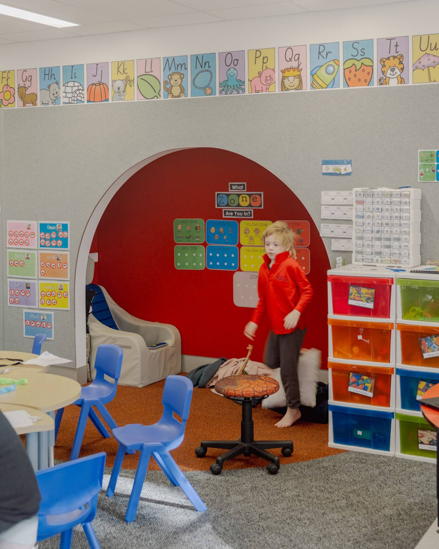 A classroom in Swan Hill Specialist School. There is a cut-out nook in a wall with pillows on the ground and an armchair. There are illustrations with the alphabet along the wall and lots of other posters. There are shelves with colourful drawers. 