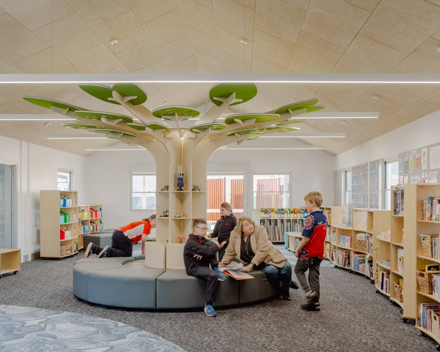 A room inside Swan Hill Specialist School that looks like a library. There are many shelves filled with books along the walls. In the middle is a seating area with a tree made out of shelves reaching to the ceiling. A teacher sits with kids reading a book. 