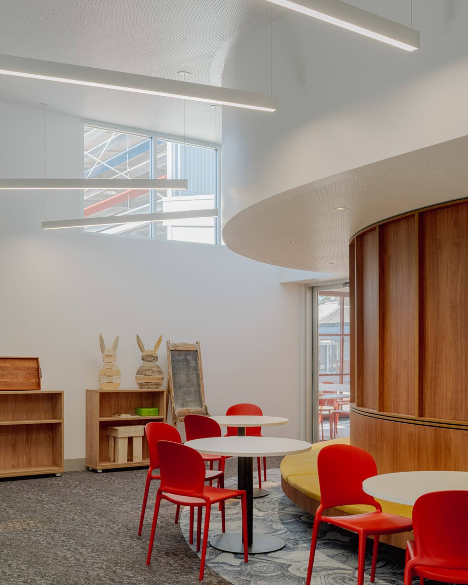 Inside Swan Hill Specialist School. There are tables with red chairs spaced around a wooden curved wall with bench seating. There are shelves in the background and long lights hanging from the ceiling. 