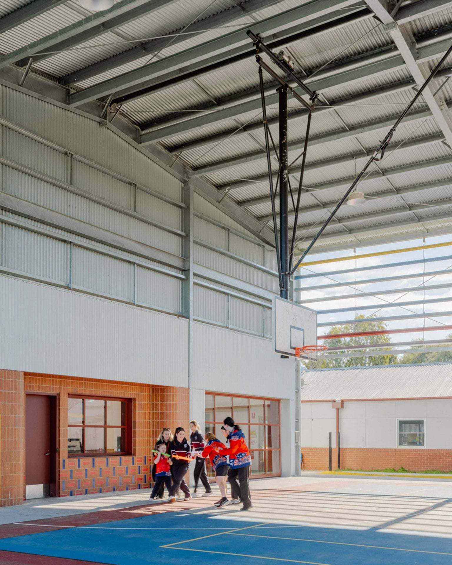 Students are playing on a hard court inside a large gymnasium. There is a basketball hoop suspended from the very high ceiling. The visible side of the gymnasium is open to the outside.