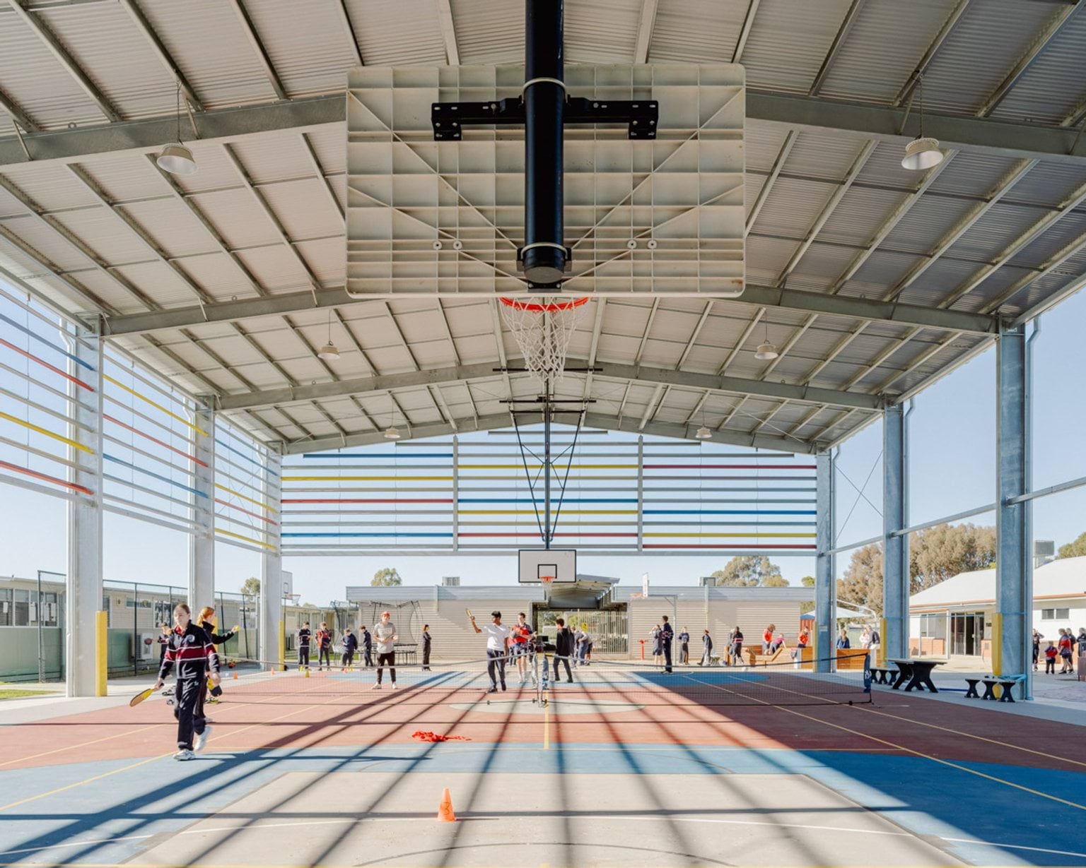 There are many students playing on a hardcourt. 3 sides of the gymnasium are open to the outside air, but the roof is covered. 2 basketball hoops are suspended from the ceiling and a tennis net is in the middle. 