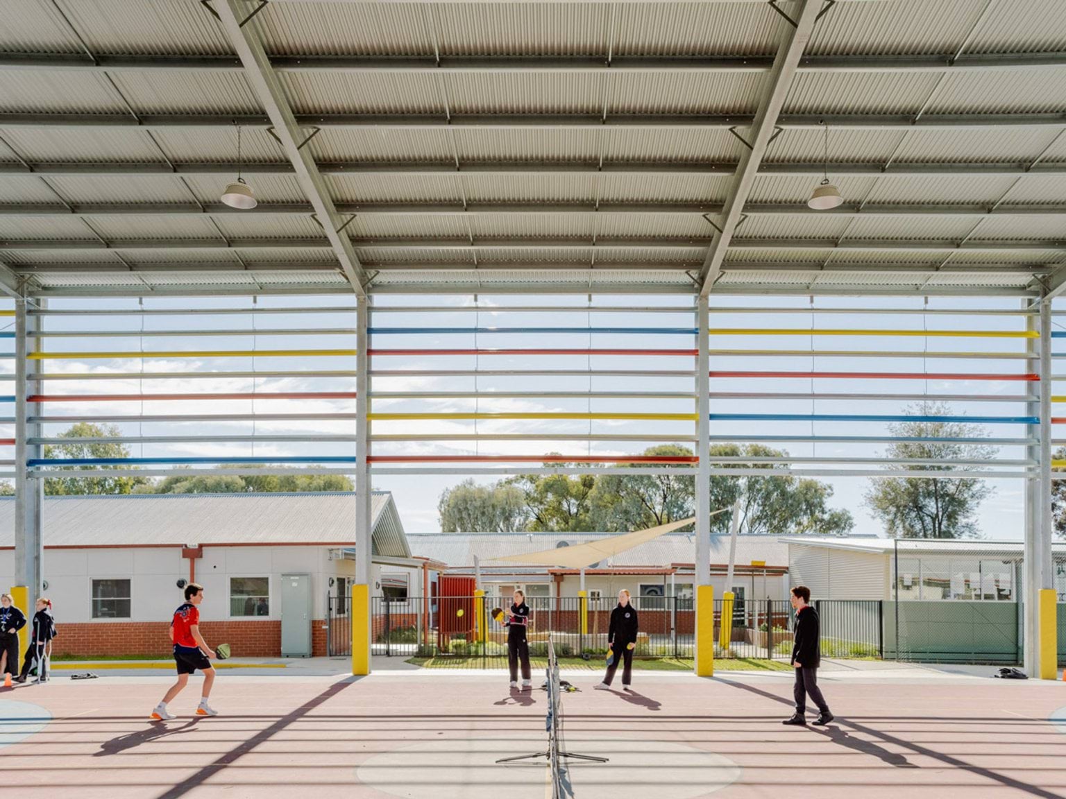 Another angle of the hard court inside the gymnaisum of Swan Hill Specialist School. The tennis net is in the middle with students on either side. The side of the gym behind them is open with coloured horizontal bars covering the top section.