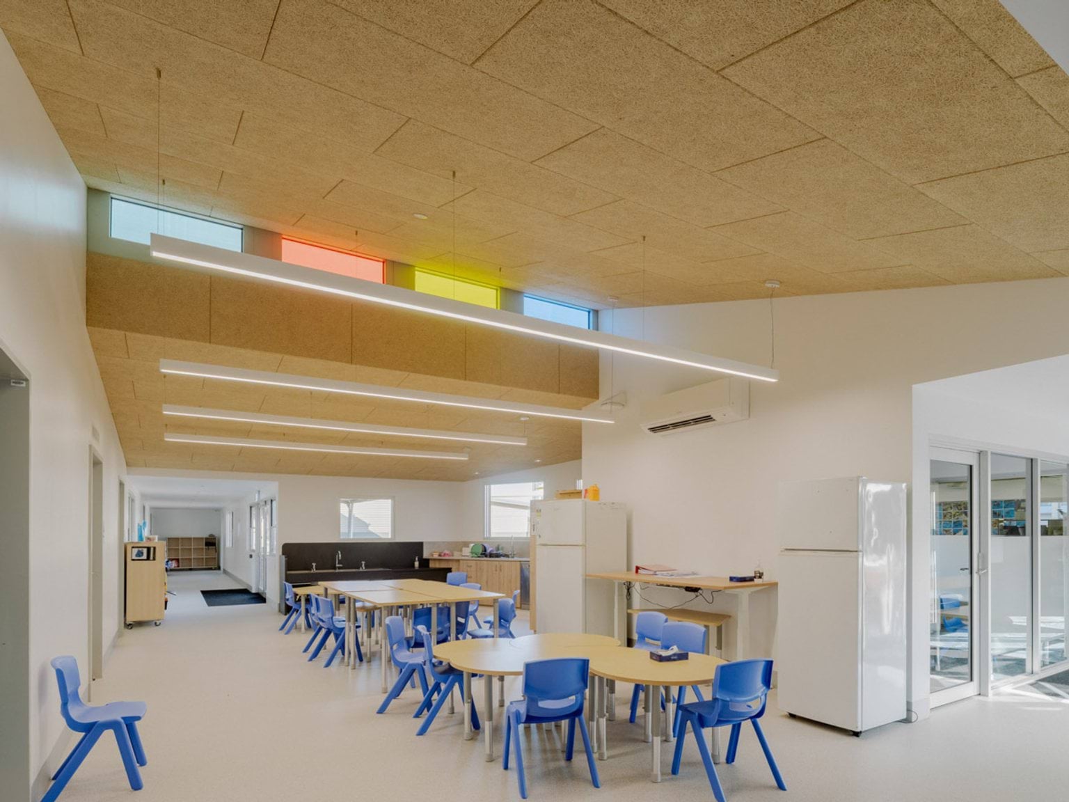 Inside a dining area of Swan Hill Specialist School. There are tables with blue chairs. There are 2 fridges along a wall.  At the top of a wall there is a row of coloured windows letting in light.