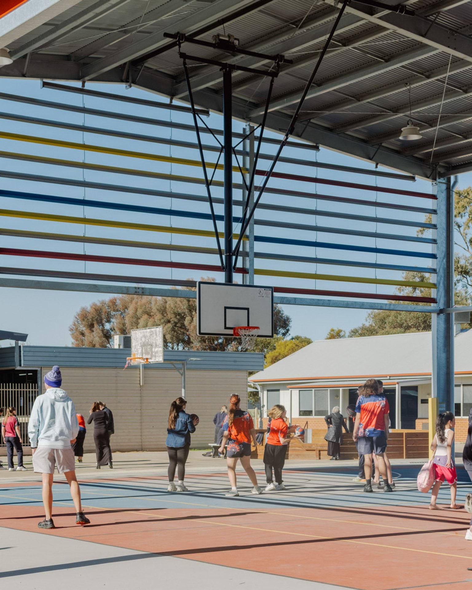Students are playing on a hard court inside a large gymnasium. There is a basketball hoop suspended from the very high ceiling. The visible side of the gymnasium is open to the outside.