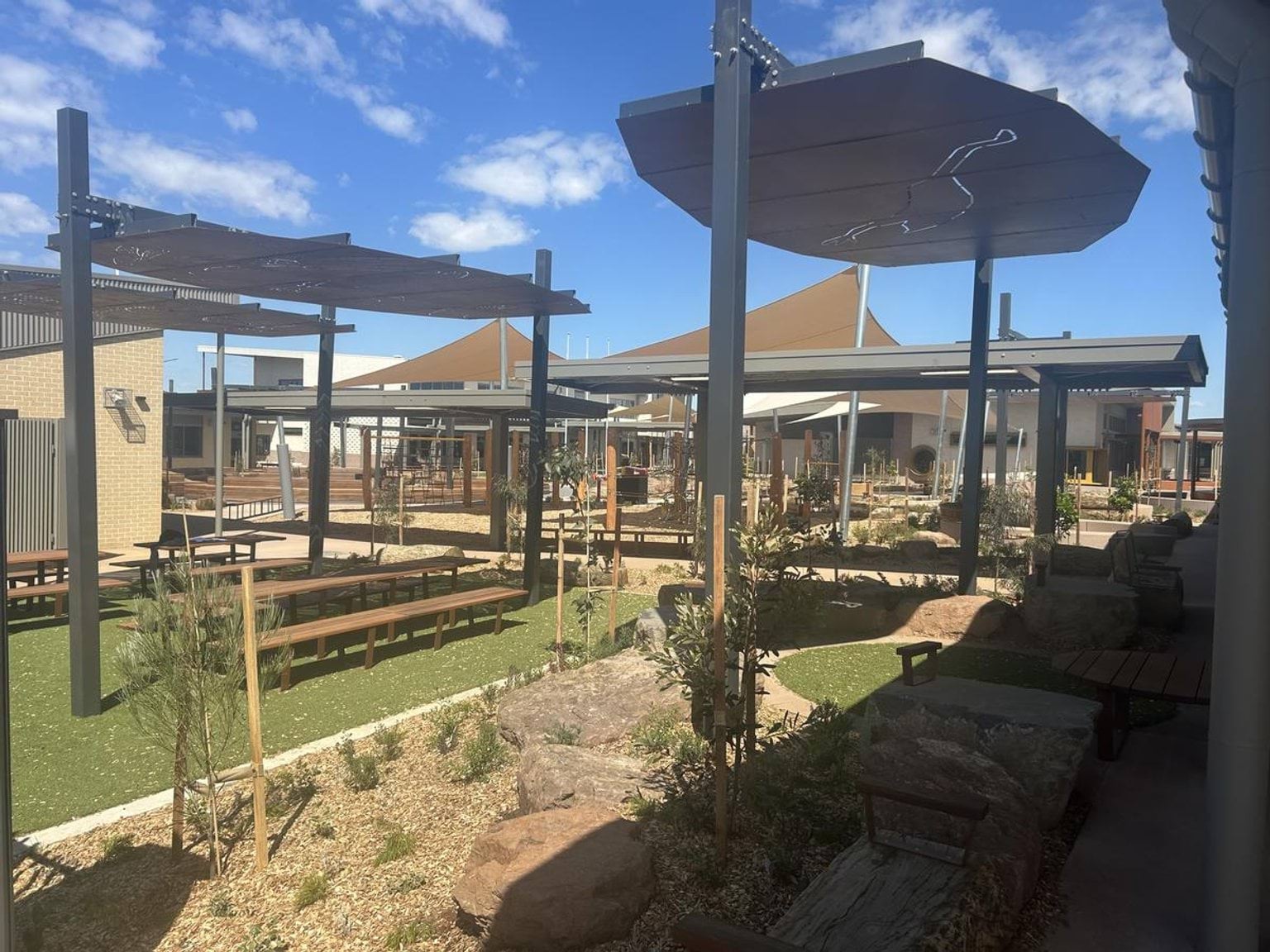 A photo of the outside area of Murrum Primary School. There are wooden tables with bench seating with shaded roofs above them. There are also new plants in the gardens.