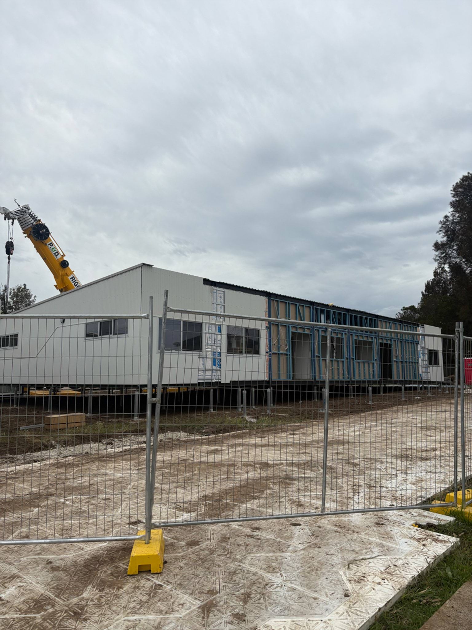 A modular building being dropped into place on the St Albans West Kindergarten site by a crane. There is a metal protective fence in front of it. 