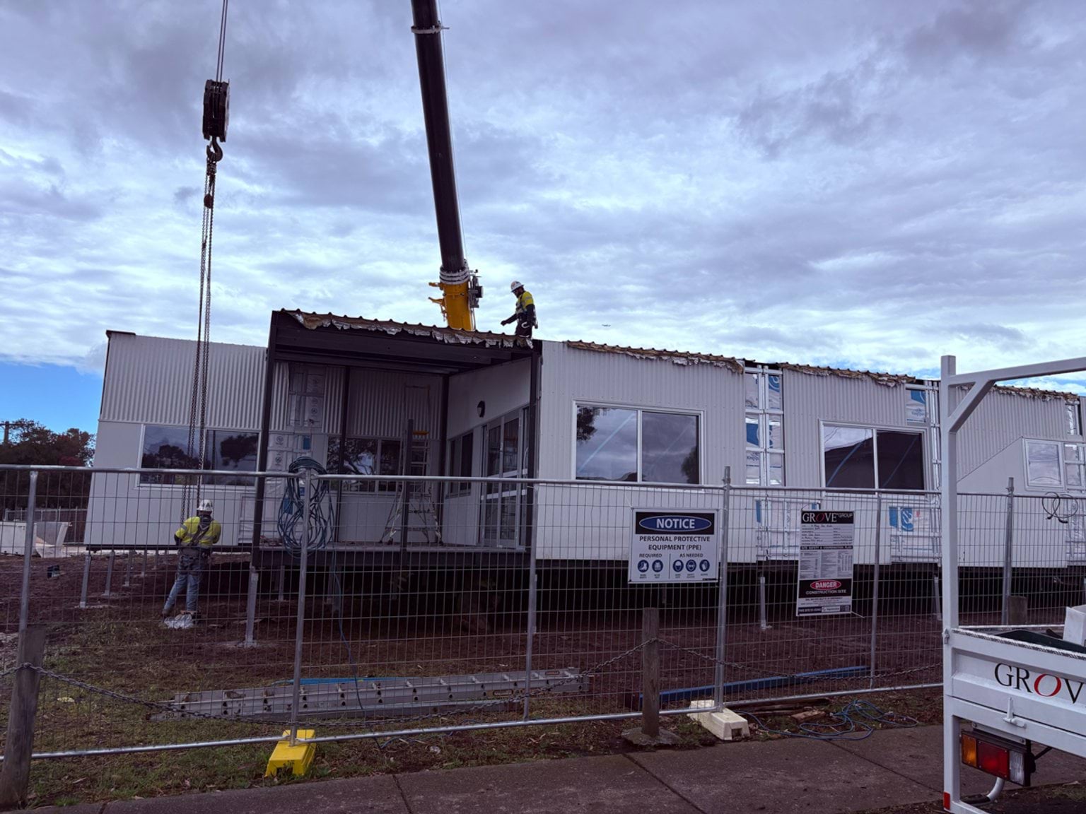 A modular building being dropped into place on the St Albans West Kindergarten site by a crane. 2 construction workers are assisting. There is a metal protective fence in front of it. 