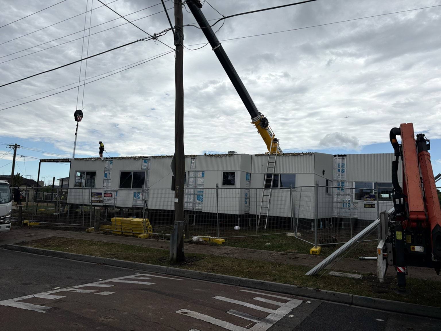 A modular building being dropped into place on the St Albans West Kindergarten site by a crane. There is a metal protective fence in front of it. A worker is on the food and there is a ladder leaning against the building.
