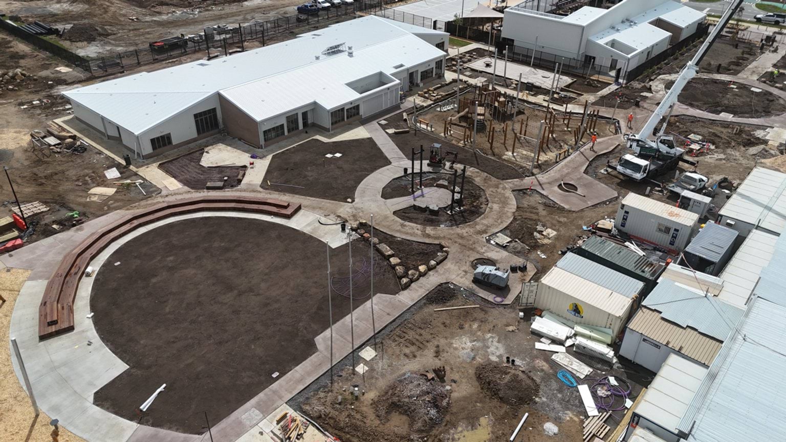 A high-angle photo of part of the outdoor area of the school. There's a bit seating area with wooden benches and freshly smoothed dirt. Concrete paths connect the area with the buildings on either side.