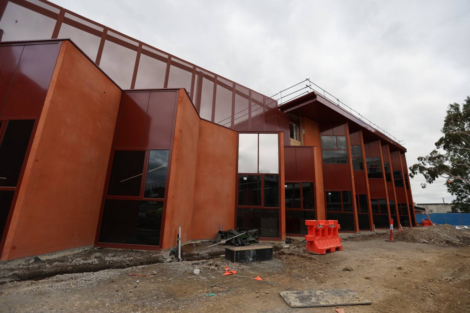 Outdoor photo of kindergarten building. It has a high roof and zigzag-style outer wall with large windows. There is construction equipment nearby.
