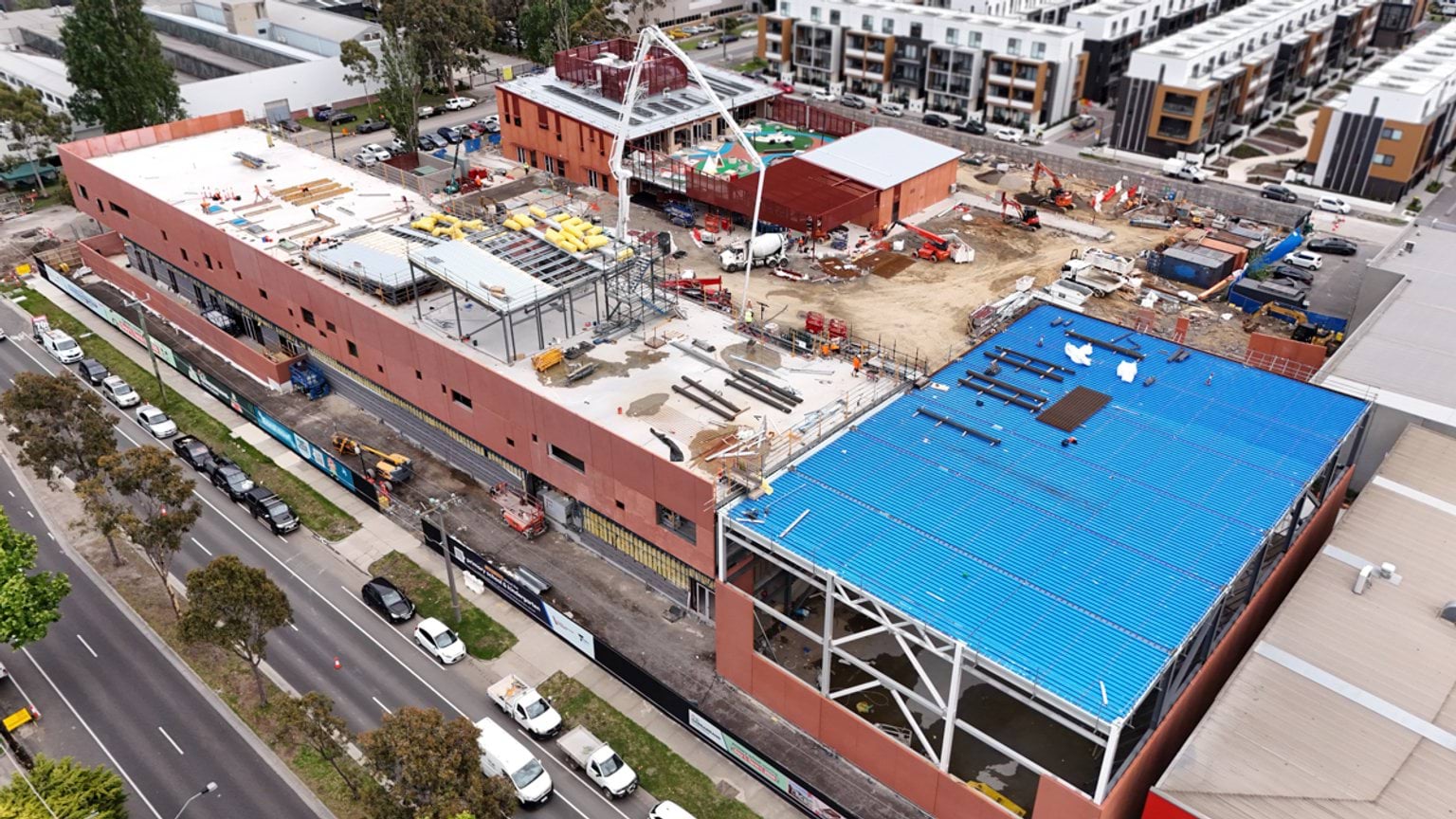 Overhead photo of kindergarten building under construction. There are multiple buildings in various stages of completion. The construction site is next to a road with parked cars.