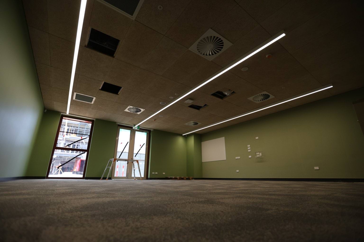 Inside of school room. There is a grey carpet, green walls, and overhead lighting with air vents, and a white board on the righthand wall. There are floor to ceililng windows at the rear.
