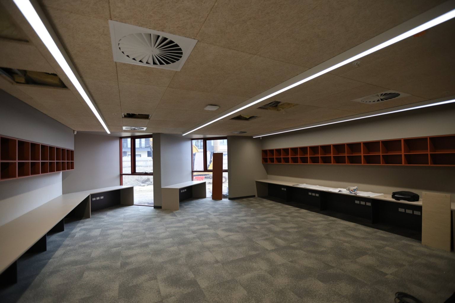 Inside view of school room. There are air vents in the ceiling, shelves and tables along the walls, and floor to ceiling windows at the rear