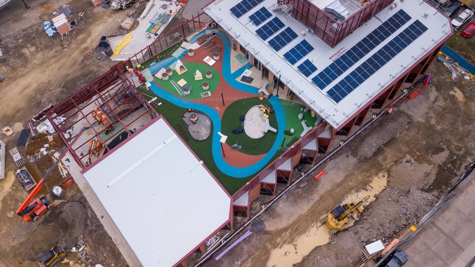 Overhead view of school under construction showing play area, school buildings with solar panels, and scaffolding