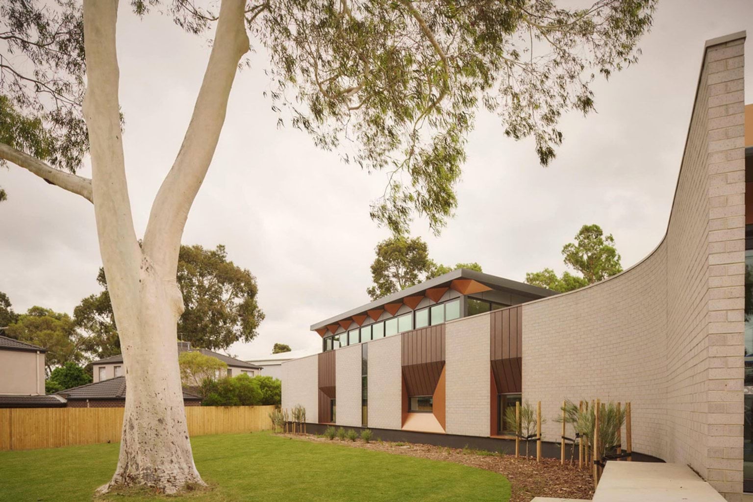 The exterior of the kindergarten with light brick walls, geometric window frames in copper tones and a curved wall. A large gum tree stands in the foreground and there is a grassed area with young plants along the building.