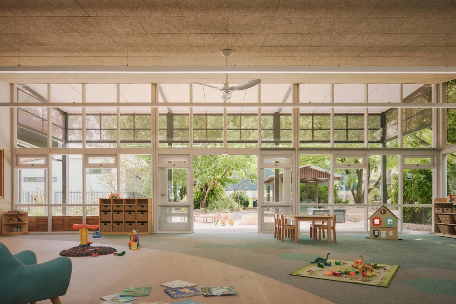 A kindergarten classroom with full-height windows opening to the outdoor play area. There are timber shelves, small tables and chairs, and toys scattered on the floor.