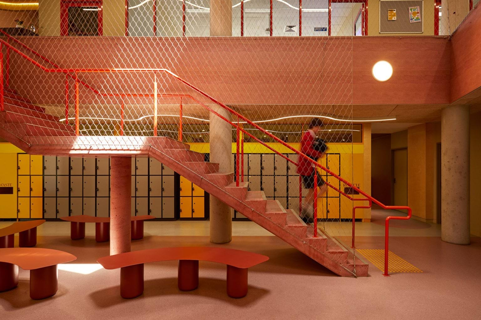 A school interior with a staircase featuring red railings and mesh panels. Yellow lockers line the walls, and benches are arranged in the open space.