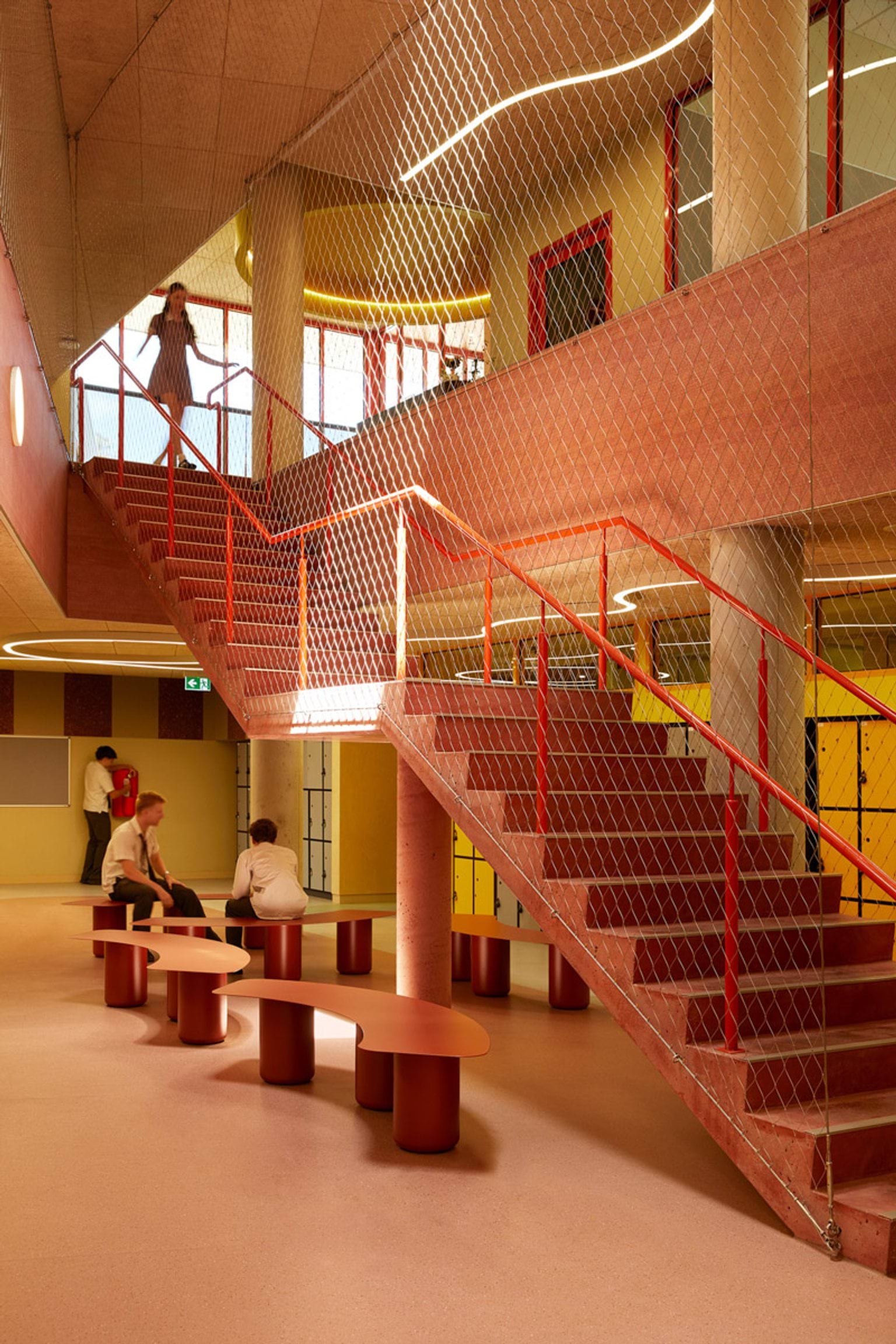 An interior staircase with red railings and mesh safety panels. The space has round tables, benches, and lockers along the walls.