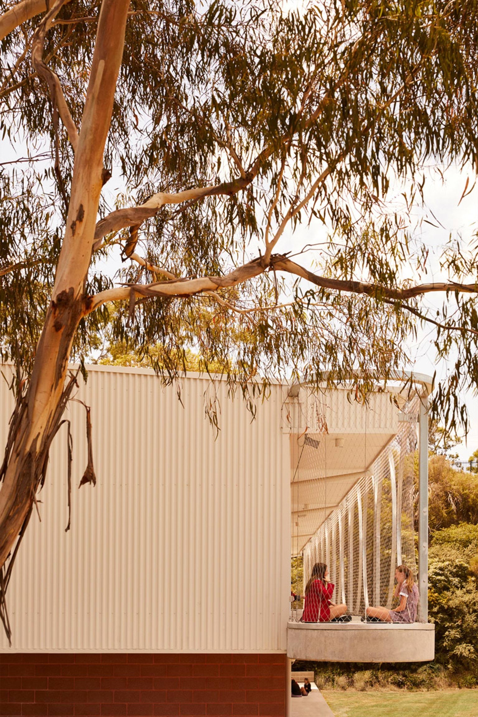 An outdoor balcony with curved metal frames and mesh panels attached to a school building. Two students are seated on the balcony, partially shaded by a tree.