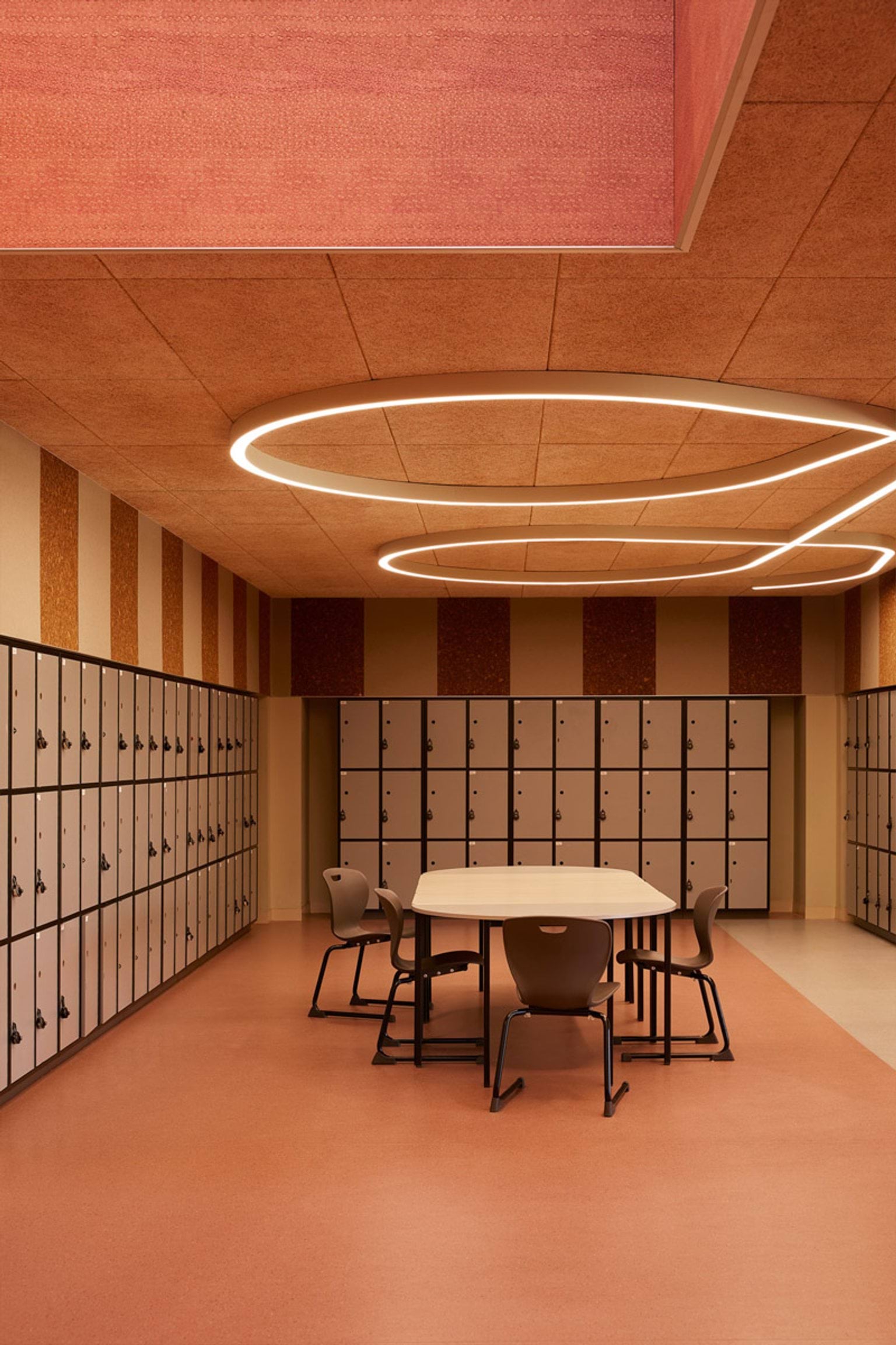 A locker area with orange flooring, striped wall panels, and circular ceiling lights. A table and chairs are placed in the centre of the space.